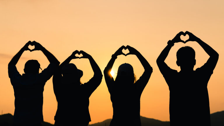 group of people with raised arms and make hand to the heart shape looking at sunrise on the mountain background